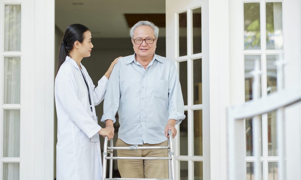 Asian senior man in eyeglasses standing with walker near the entrance of his house and trying to go out with his home caregiver